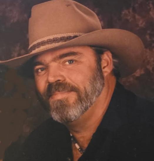 Man in cowboy hat with beard, wearing a dark shirt, posing against a neutral background.