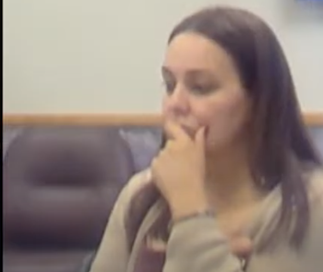 A woman in thoughtful pose during a meeting, hands near mouth, seated in a chair with blurred background.