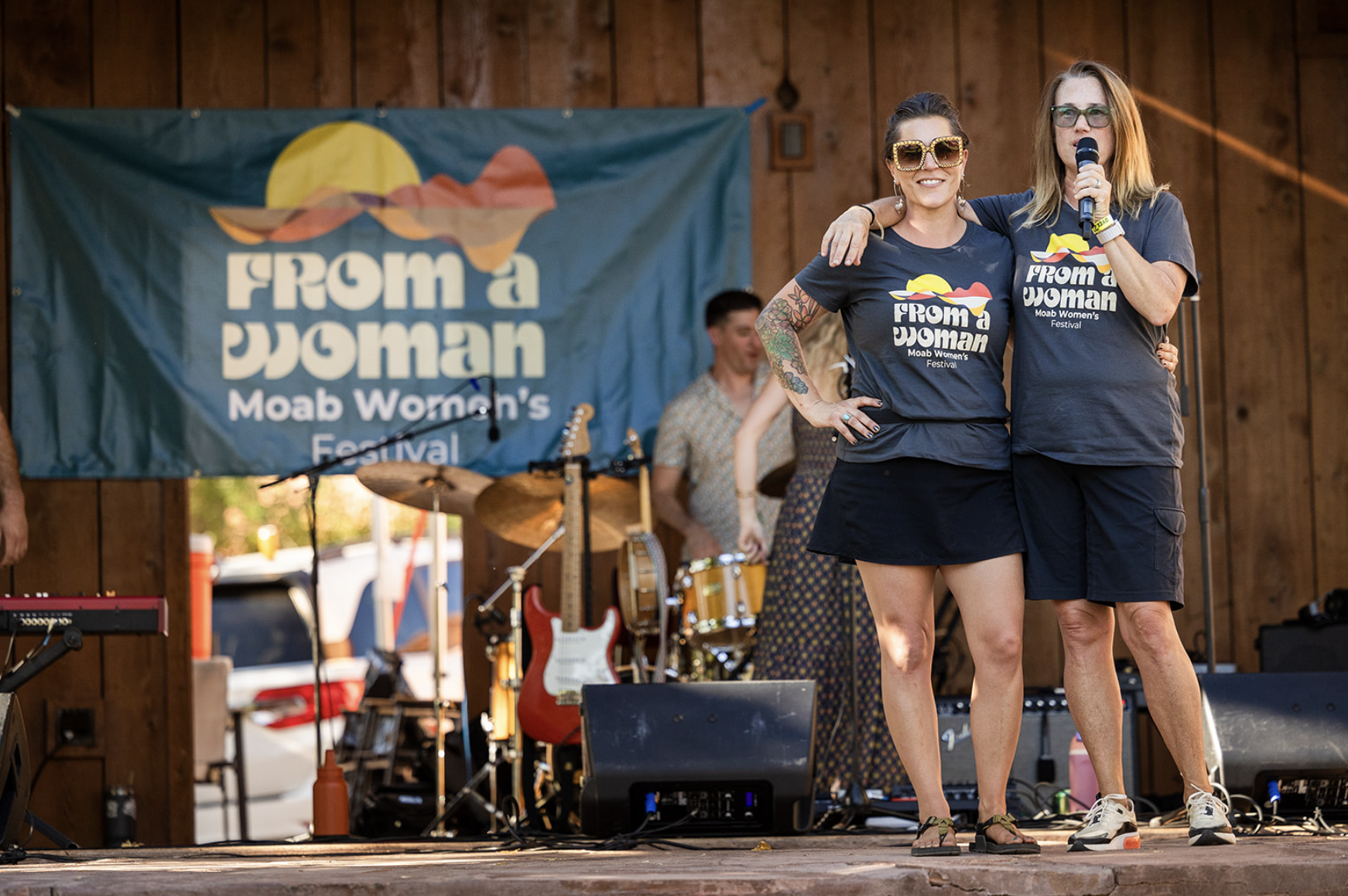 Two women on stage at Moab Women's Festival, From a Woman banner in background.