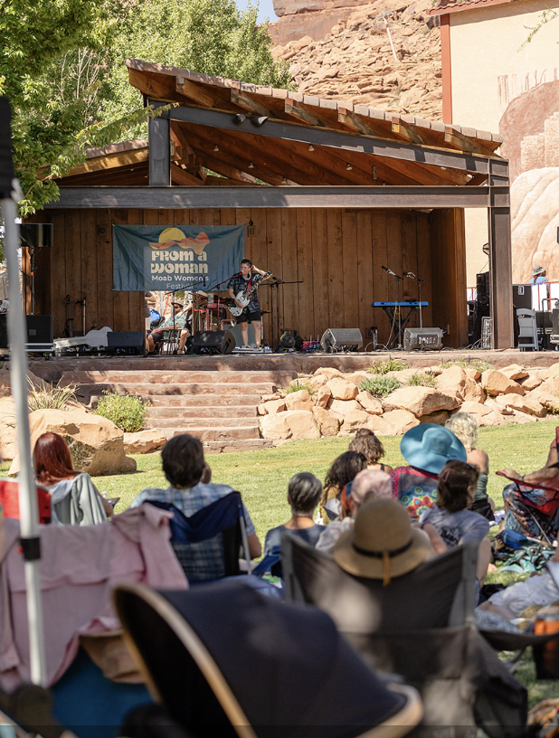 Outdoor concert at a wooden stage with audience seated on grass, featuring From a Woman Moab Women's Fest sign.