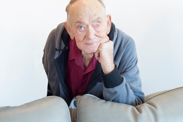 Elderly man in casual attire leaning on a couch, gazing thoughtfully at the camera against a white background.