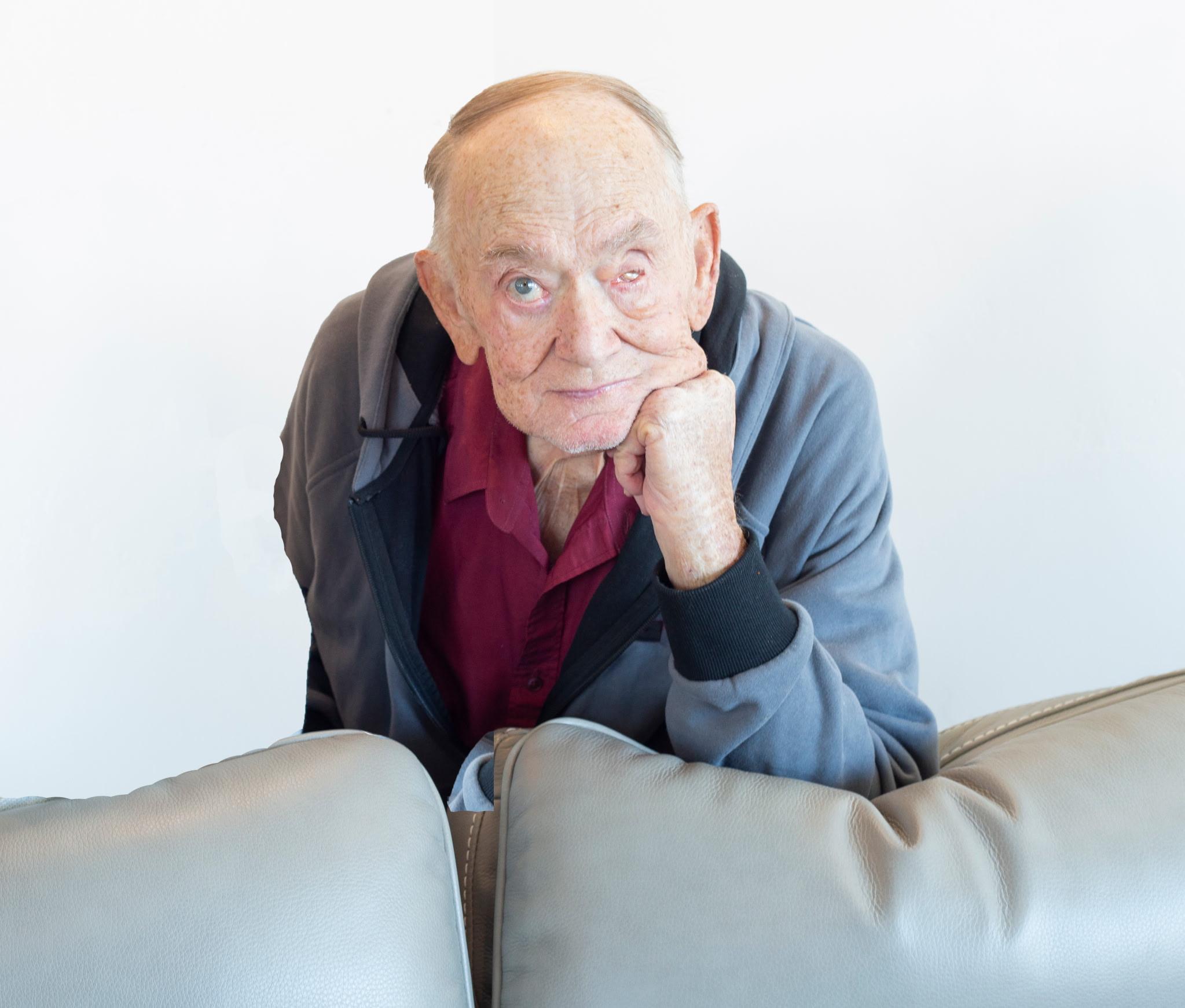 Elderly man in casual attire leaning on a couch, gazing thoughtfully at the camera against a white background.