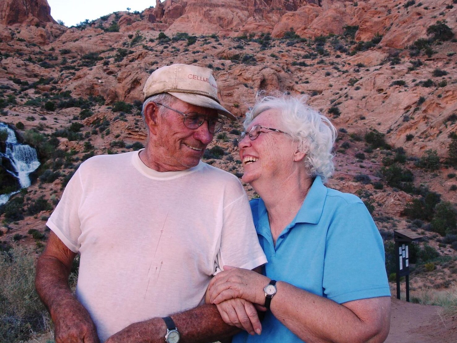 Couple smiling at desert landscape with rocky hills and waterfall in the background.