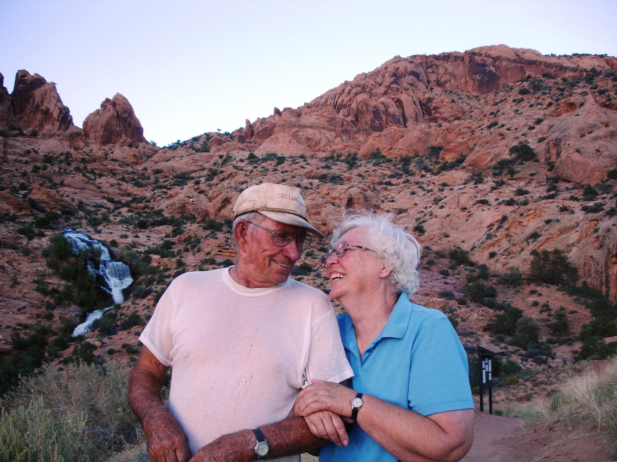 Couple smiling at desert landscape with rocky hills and waterfall in the background.