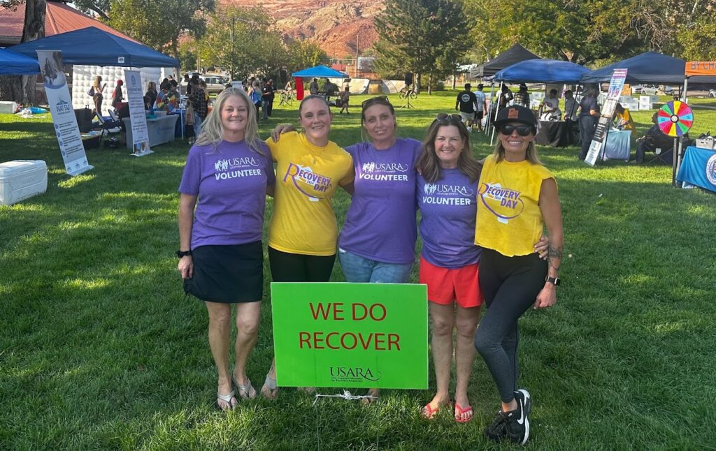 Volunteers at an outdoor recovery event with a We Do Recover sign, surrounded by booths and trees.