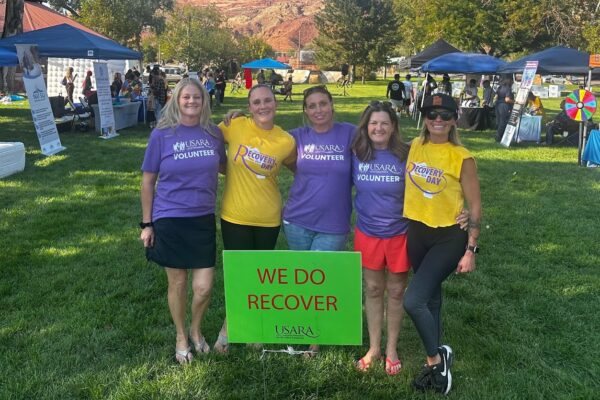 Volunteers at an outdoor recovery event with a We Do Recover sign, surrounded by booths and trees.
