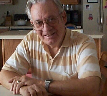 Older man with glasses smiles, wearing a striped polo shirt, sitting at a kitchen table in a cozy home setting.