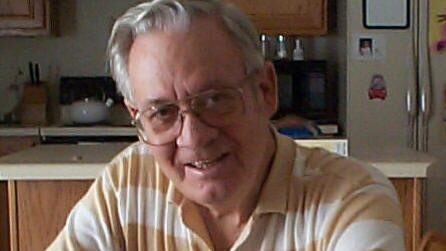 Older man with glasses smiles, wearing a striped polo shirt, sitting at a kitchen table in a cozy home setting.