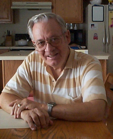 Older man with glasses smiles, wearing a striped polo shirt, sitting at a kitchen table in a cozy home setting.