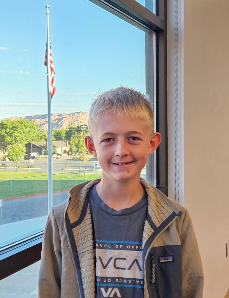 Young boy smiling indoors with an American flag and landscape visible outside through the window.