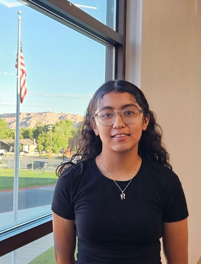 Young woman with curly hair in front of a window with an American flag outside and hills in the background.