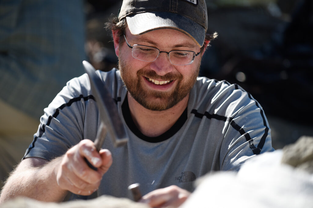 Smiling man using a hammer, wearing glasses and a cap, engaged in outdoor activity.