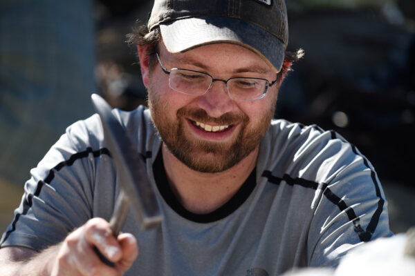 Paleontologist Randall Irmis using a hammer, wearing glasses and a cap, engaged in outdoor activity.