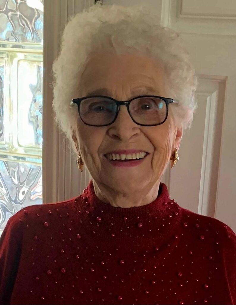 Elderly woman smiling in glasses and a red sweater with pearls, standing indoors by a textured glass window.
