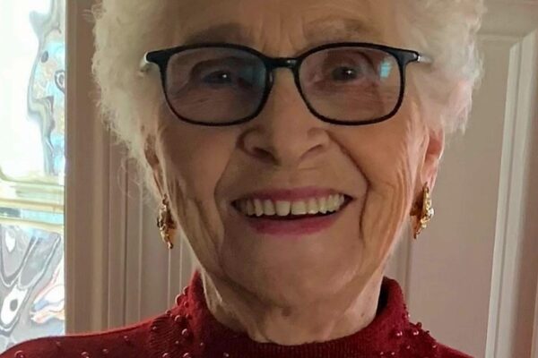 Elderly woman smiling in glasses and a red sweater with pearls, standing indoors by a textured glass window.