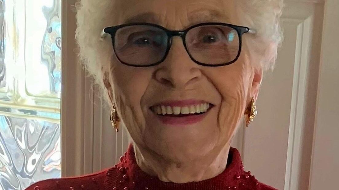 Elderly woman smiling in glasses and a red sweater with pearls, standing indoors by a textured glass window.