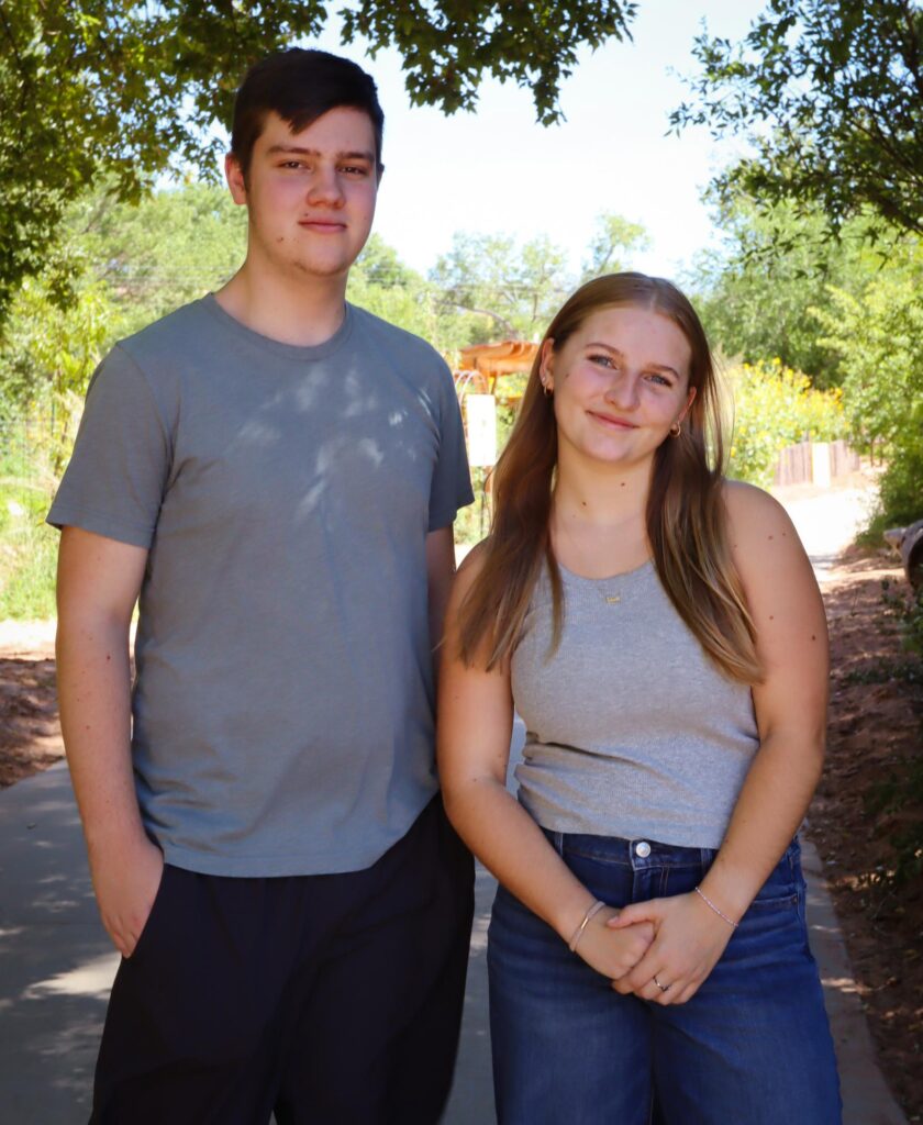 Two people smiling outdoors on a sunny day, standing on a pathway surrounded by trees and greenery.