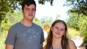 Two people smiling outdoors on a sunny day, standing on a pathway surrounded by trees and greenery.