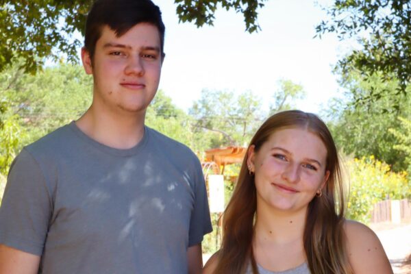 Two people smiling outdoors on a sunny day, standing on a pathway surrounded by trees and greenery.