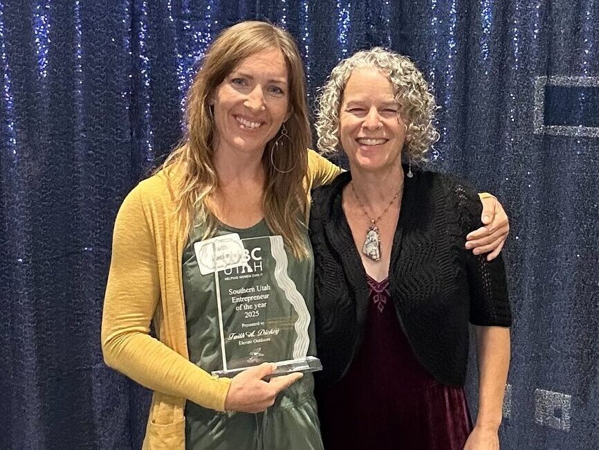 Two women smiling, one holding an award, standing in front of a blue curtain at a formal event.