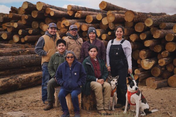 Group of people and dogs posing in front of stacked logs outdoors, wearing warm clothing.