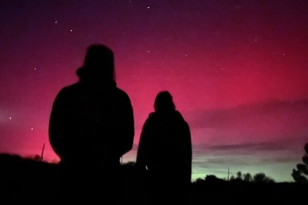 People observing the Aurora Borealis at Moab's Sandflats Recreation Area. Silhouettes under a vibrant pink and purple night sky, possibly observing aurora borealis.