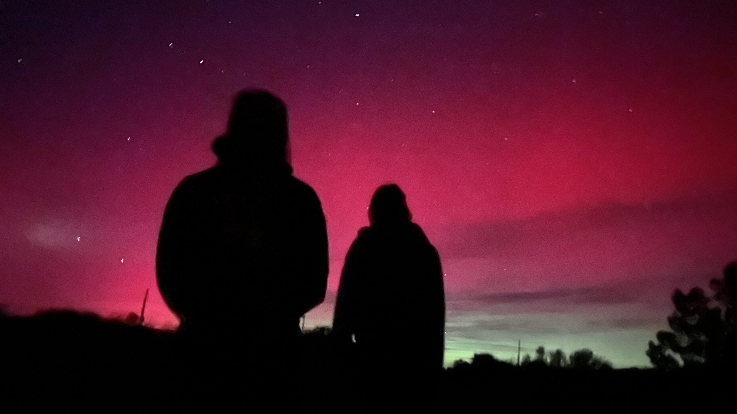 People observing the Aurora Borealis at Moab's Sandflats Recreation Area. Silhouettes under a vibrant pink and purple night sky, possibly observing aurora borealis.
