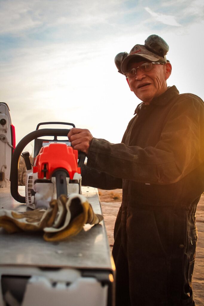 Man in safety gear handling a chainsaw outdoors at sunset, wearing ear protection and gloves nearby.Warm Elders