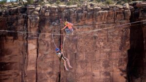Two people highlining across a canyon, one hanging upside down, showcasing extreme adventure and balance skills. Suspended above a Moab canyon, Faith Dickey with Elevate Outdoors helps veterans with 22 Jumps confront fear, trust their safety systems, and step into the void. Through patience, breath, and compassion, fear becomes a teacher—not an enemy—on the Skywalk.