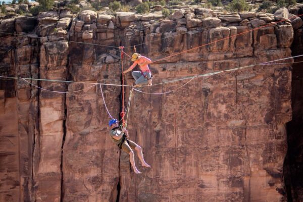 Two people highlining across a canyon, one hanging upside down, showcasing extreme adventure and balance skills. Suspended above a Moab canyon, Faith Dickey with Elevate Outdoors helps veterans with 22 Jumps confront fear, trust their safety systems, and step into the void. Through patience, breath, and compassion, fear becomes a teacher—not an enemy—on the Skywalk.