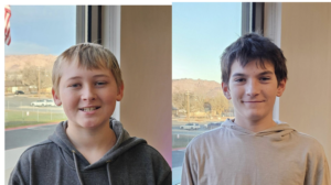 Two smiling boys in hoodies standing indoors, with a window view of a scenic outdoor landscape in the background.