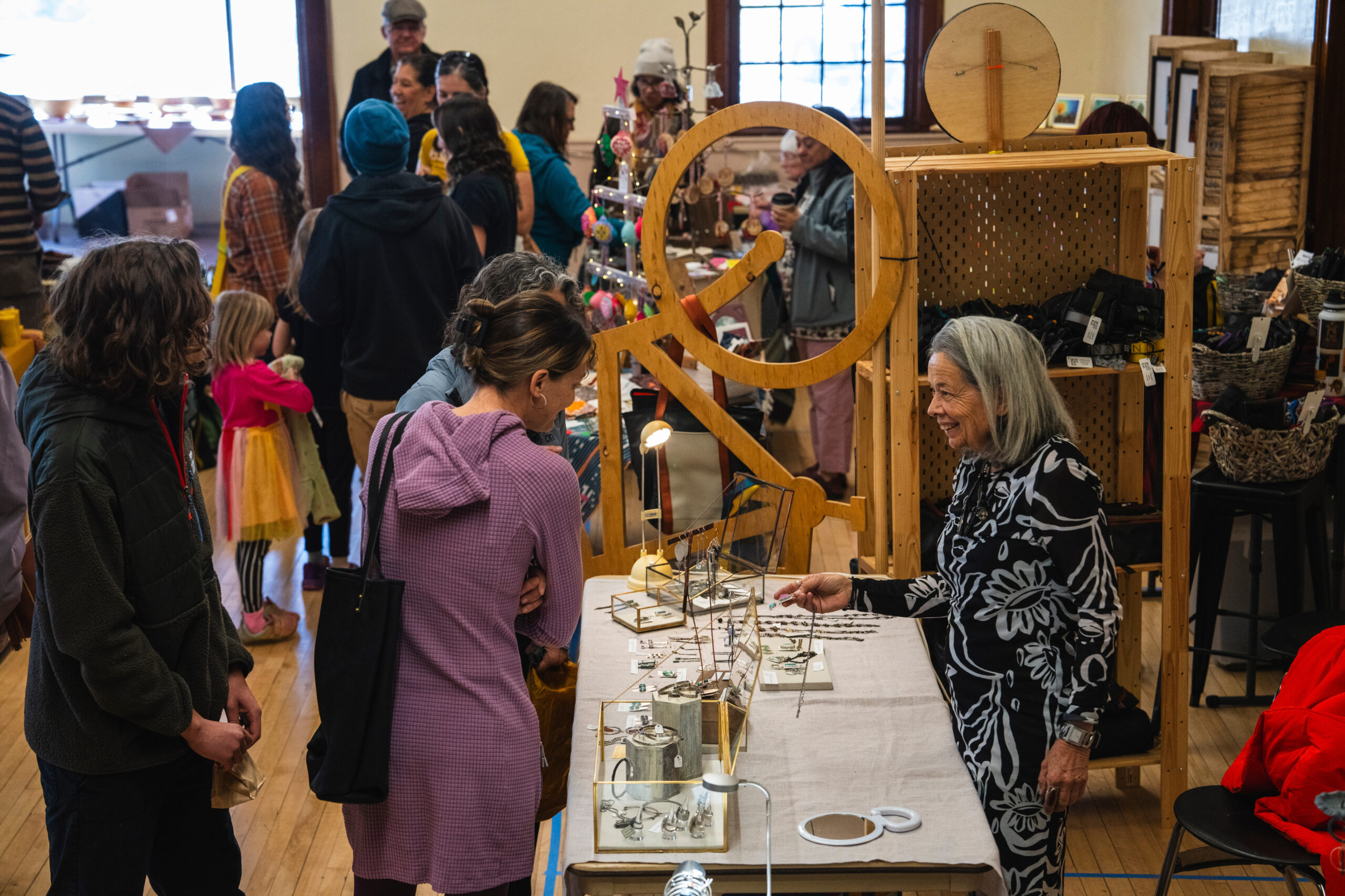 A joyful woman sells handmade jewelry at a bustling craft fair, surrounded by interested customers and vibrant displays.