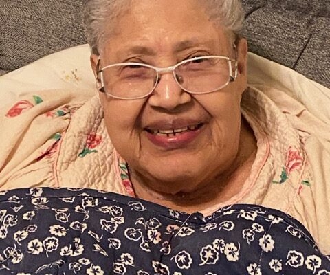 Elderly woman smiling warmly with glasses, wrapped in a floral blanket, sitting comfortably indoors.