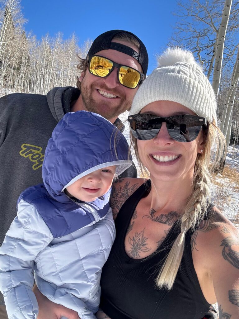Hill Family enjoying a sunny winter day outdoors, smiling and bundled up in warm clothing with snowy trees in the background.