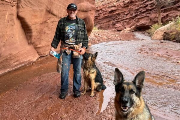 Man hiking with two German Shepherds, one who became a lost dog outside Moab, in a scenic red rock canyon with a small stream and autumn trees.