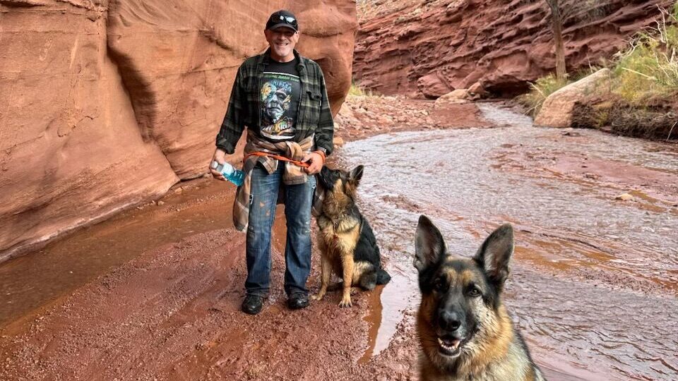 Man hiking with two German Shepherds, one who became a lost dog outside Moab, in a scenic red rock canyon with a small stream and autumn trees.