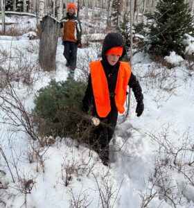 Boy in orange vest dragging a small Christmas tree through snowy forest, followed by another person. Moab's Gustafson Family celebrating Christmas