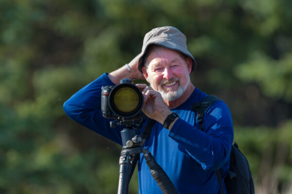 Elderly man with camera on tripod, smiling outdoors in blue shirt and hat, enjoying photography.