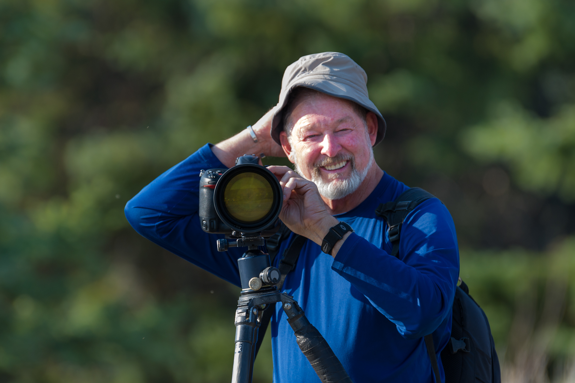 Elderly man with camera on tripod, smiling outdoors in blue shirt and hat, enjoying photography.