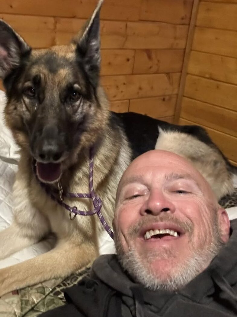 Man and German Shepherd smiling together on a bed in a cozy wooden room.