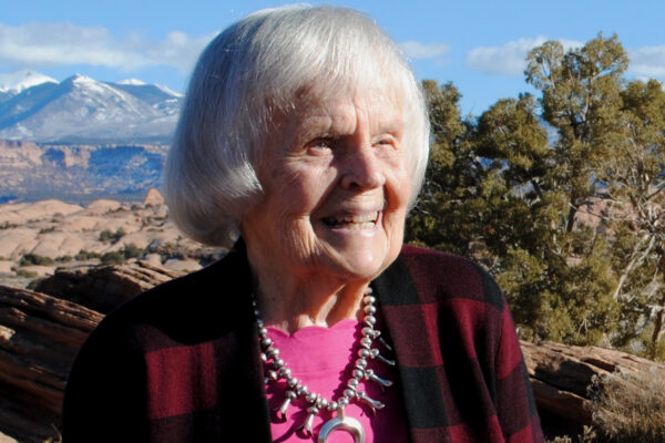 Elderly woman smiling outdoors in rugged landscape with mountains and trees under clear blue sky.