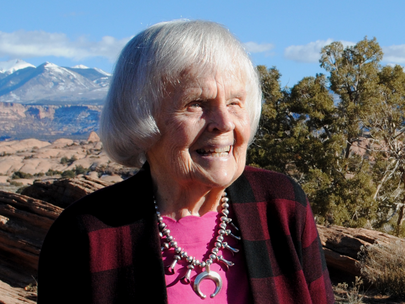 Elderly woman smiling outdoors in rugged landscape with mountains and trees under clear blue sky.