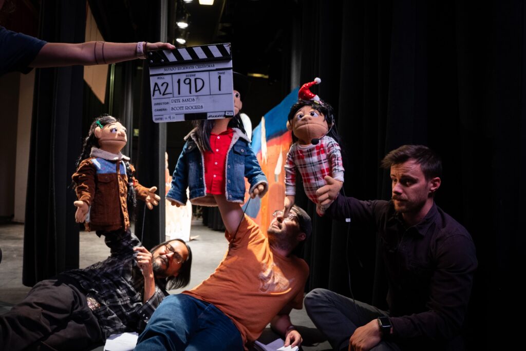 Puppeteers arranging puppets on a theater set under stage lights with a clapperboard visible in the background.