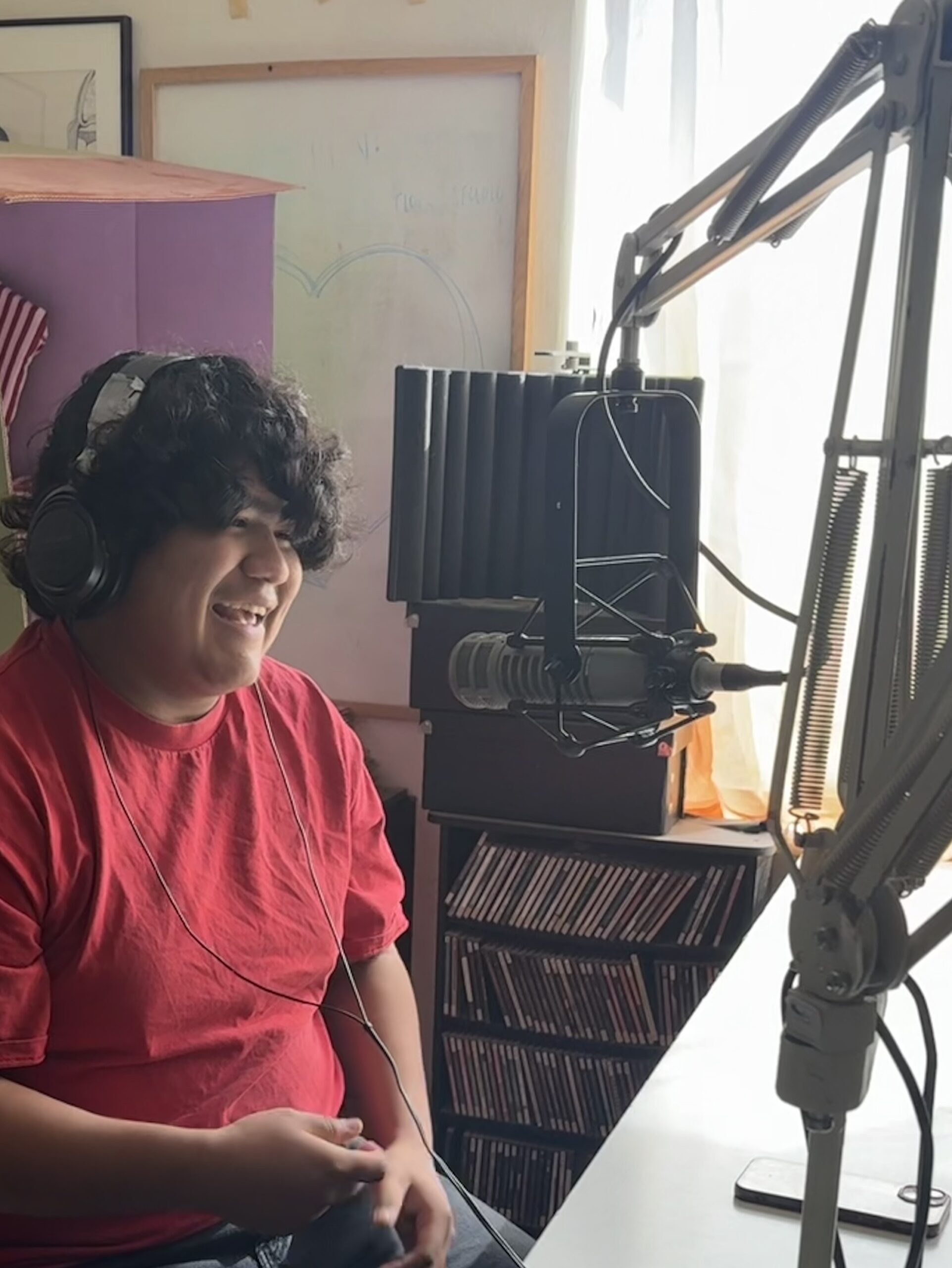 Person recording a podcast in a studio, wearing headphones and smiling, with a microphone and sound equipment.