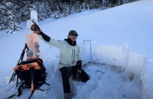 Utah Avalanche Center worker analyzes snowpack with skis and gear in snowy forest, pointing left.