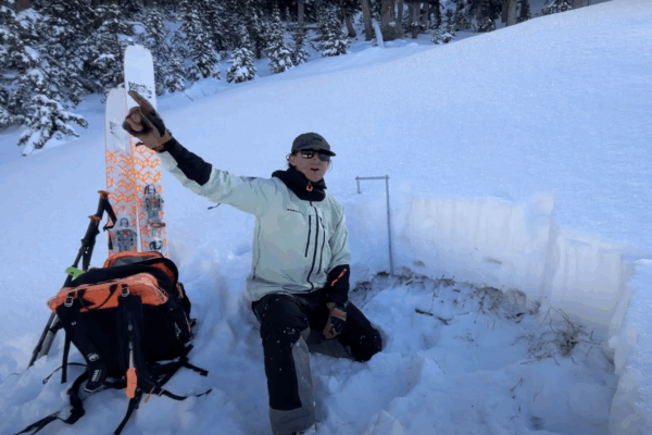 Utah Avalanche Center worker analyzes snowpack with skis and gear in snowy forest, pointing left.
