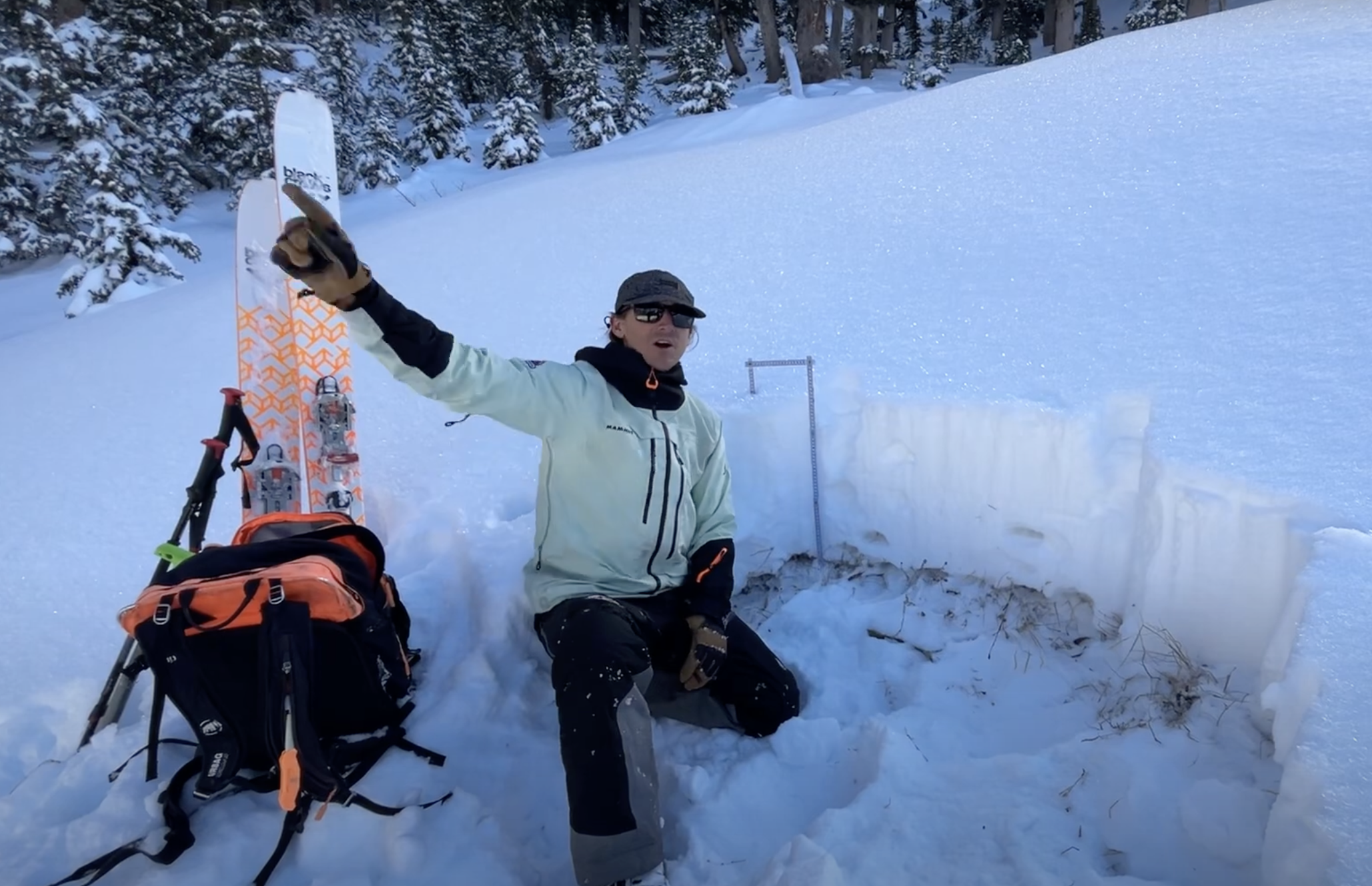 Utah Avalanche Center worker analyzes snowpack with skis and gear in snowy forest, pointing left.