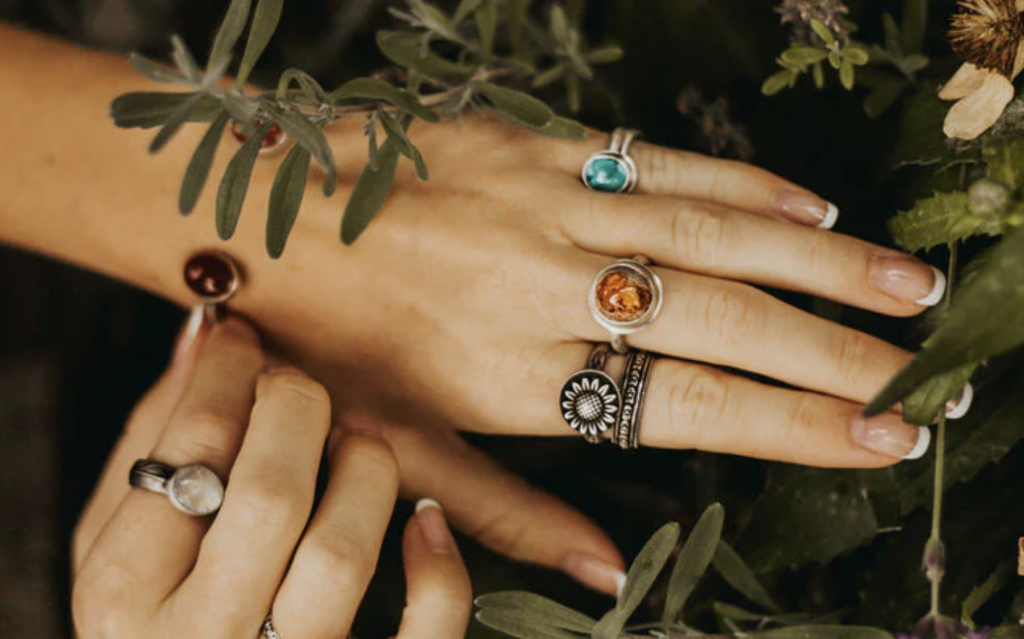 Hands adorned with colorful gemstone rings amidst green foliage.