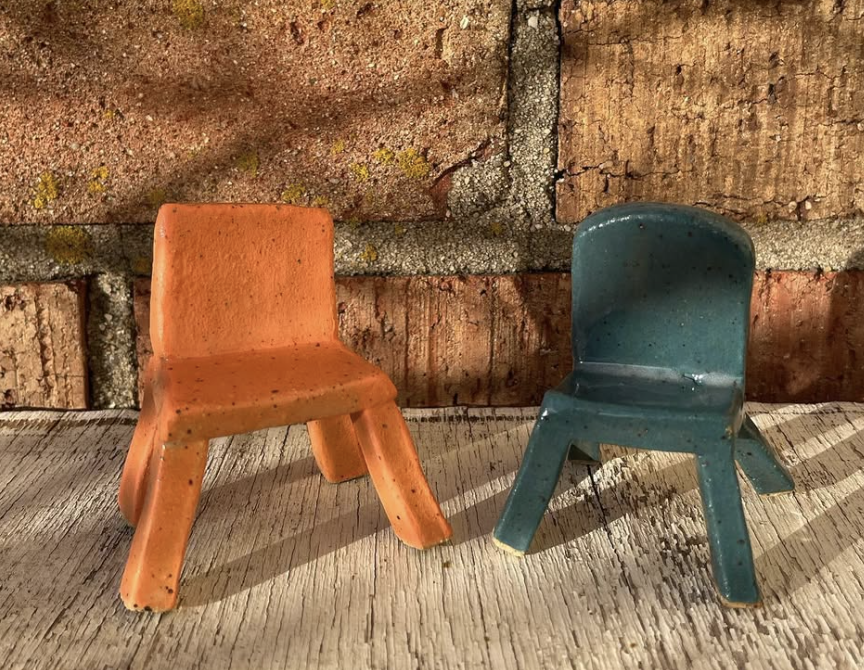 Two small clay chairs, one orange and one blue, on a wooden surface with a brick wall backdrop.