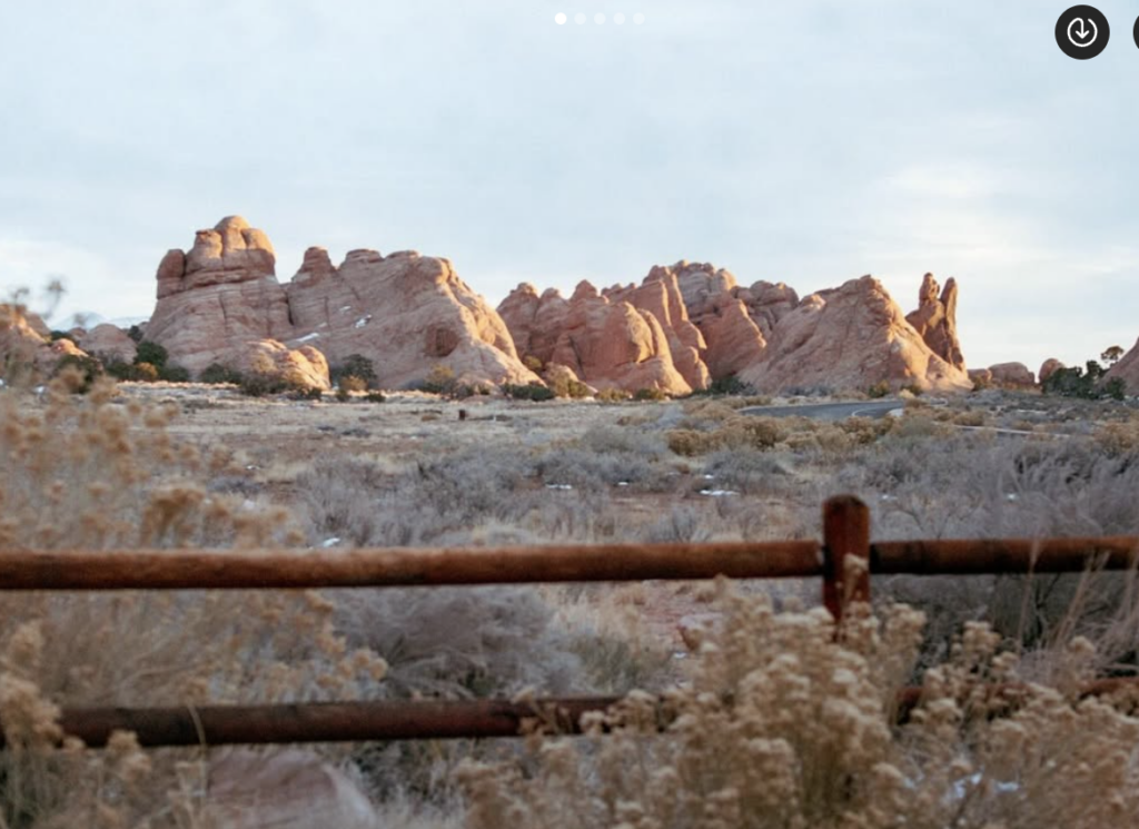 Red rock formations at sunrise seen beyond a rustic wooden fence, with a dry landscape in the foreground.